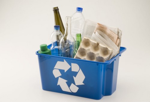A blue plastic recycling bin positioned on a plain light-colored background, filled with various household waste items. Inside the bin, there are clear glass bottles, including a small green plastic bottle, a larger brown glass bottle, and a clear glass bottle with a blue cap. Additionally, there is a transparent plastic water bottle with a blue cap, as well as a cardboard egg carton and crumpled paper with printed text. The waste materials occupy most of the binâ€™s interior, with some items leaning slightly against the sides. The recycling symbol printed in white on the front of the bin signifies its intended use for recyclable rubbish. The scene suggests a typical collection area for waste removal services, such as those provided by Commercial Waste Removal Gipsy Hill, serving local businesses or residential settings within the Gipsy Hill postcode area, emphasizing environmentally responsible waste disposal.