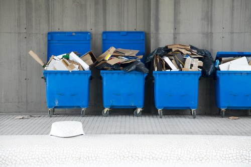A row of four blue wheeled commercial rubbish bins positioned on a paved surface along a concrete wall, with the first bin on the left partially overflowing with mixed waste, including white paper, cardboard, and other lightweight packaging materials. The second bin is also filled with various cardboard boxes and paper debris, some of which are leaning against the sides and spilling over the top. The third bin appears to contain similar waste, with a black plastic bag partially open revealing more paper and cardboard inside. The fourth bin, on the far right, is less filled or less visible. In front of the bins, on the pavement, lies a loose piece of white paper or cardboard, while the background features a plain, grey concrete wall. The setting suggests a commercial or industrial area, typical for waste collection and rubbish removal services in Gipsy Hill, London, undertaken by Commercial Waste Removal Gipsy Hill, serving local rubbish disposal needs.