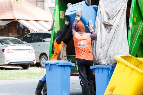 Two waste collection workers, wearing orange high-visibility vests and gloves, are lifting a large blue plastic bin toward the hopper of a green rubbish disposal vehicle, which is parked on a street in a residential area. The workers are standing on the roadside pavement with three other empty blue bins and one yellow bin positioned nearby for collection. In the background, a silver car is parked facing away from the scene, and a brick building with a striped awning and trees are visible, indicating a suburban environment. The scene appears to be during daytime, with natural light illuminating the workers and waste equipment, emphasizing the practical nature of rubbish removal services in the Gipsy Hill area, suggesting a routine waste collection process typical in urban and suburban settings around London.