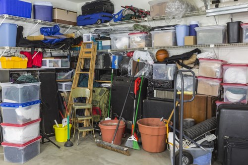 A cluttered storage area or garage interior filled with various plastic storage boxes, some transparent and others opaque, stacked on metal shelving units. The boxes are in multiple colours including blue, black, white, and red, and contain miscellaneous items, with some lids off or partially open. In the foreground, there are two large terracotta plant pots, tipped over and surrounded by cleaning tools such as brooms, mops, and a broomstick, leaning against a stack of chairs and a hand trolley. A small yellow bucket and a black hand truck are also present among the organized clutter. The shelving unit extends across the background, holding additional containers, cardboard boxes, and various household or workshop items in an indoor environment typical of a residential garage or storage space. Commercial Waste Removal Gipsy Hill occasionally handles such clutter for rubbish removal services in the Gipsy Hill area, made evident by the dense accumulation of miscellaneous household waste and storage items ready for collection.