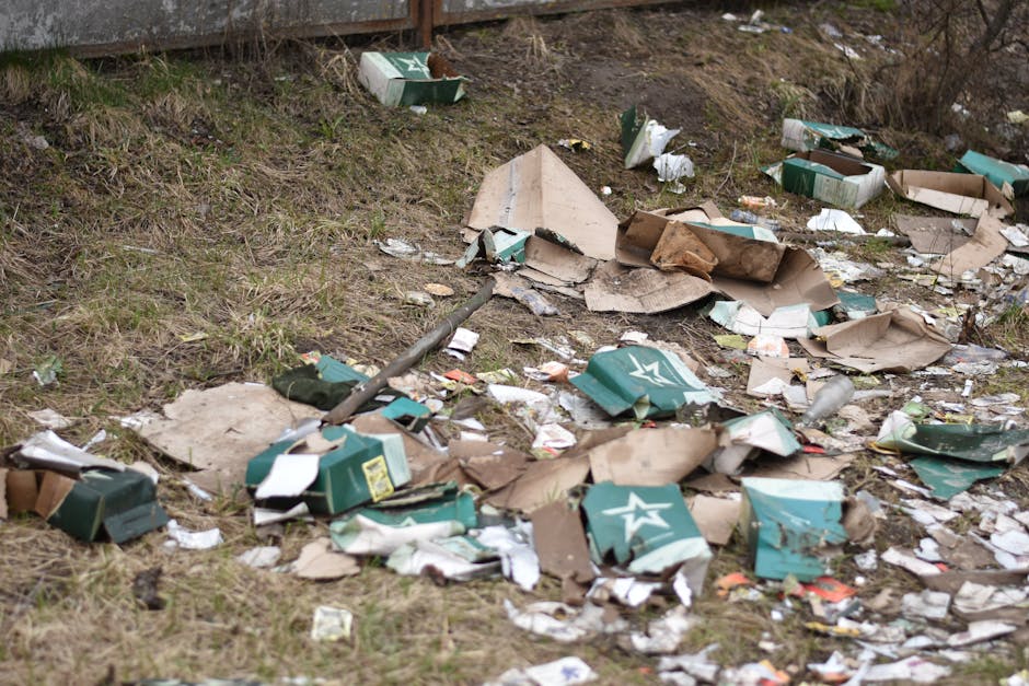 On a paved sidewalk next to a black metal fence with pointed finials and dense foliage in the background, three large black plastic garbage bags filled with waste are leaning against the fence. The ba